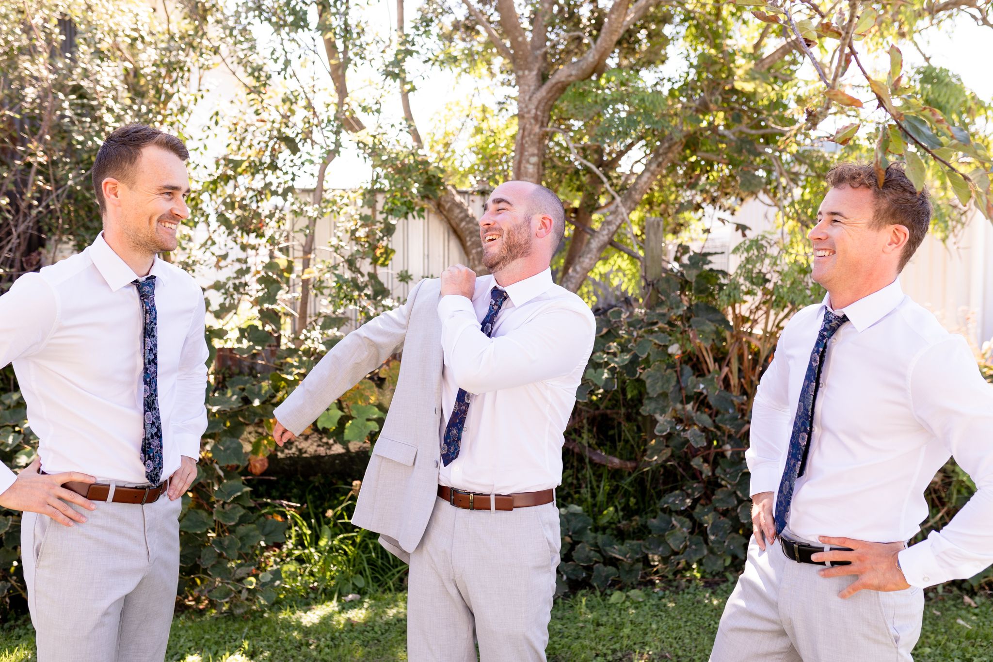 Three people in white shirts and ties share a lighthearted moment in a garden setting.