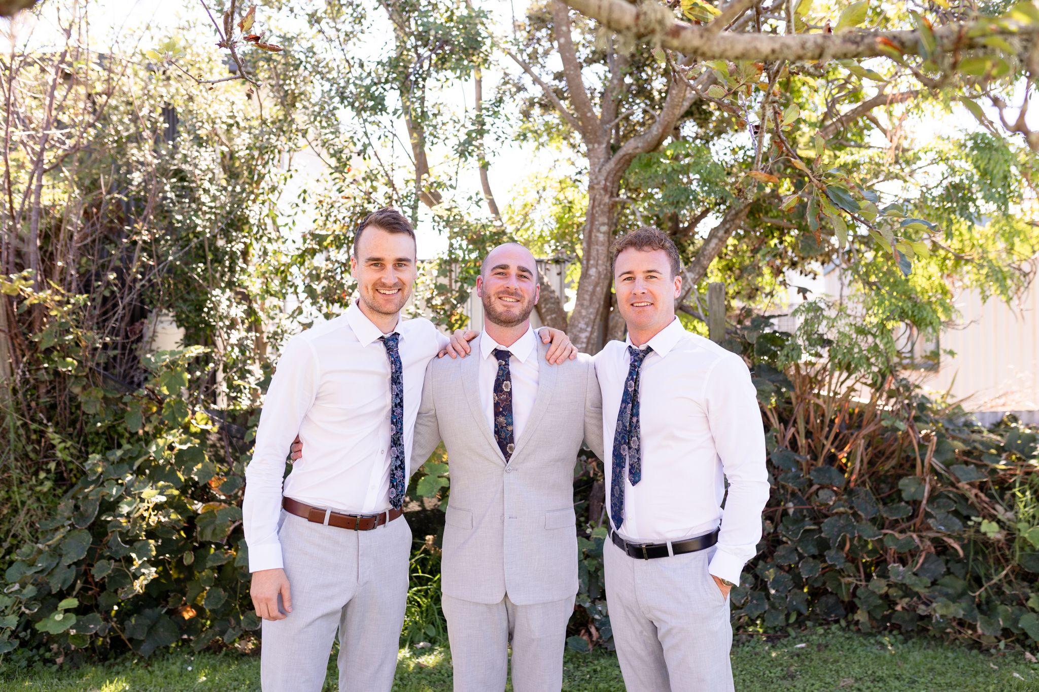 Three people in coordinated white and light-colored formal attire pose together in a garden.