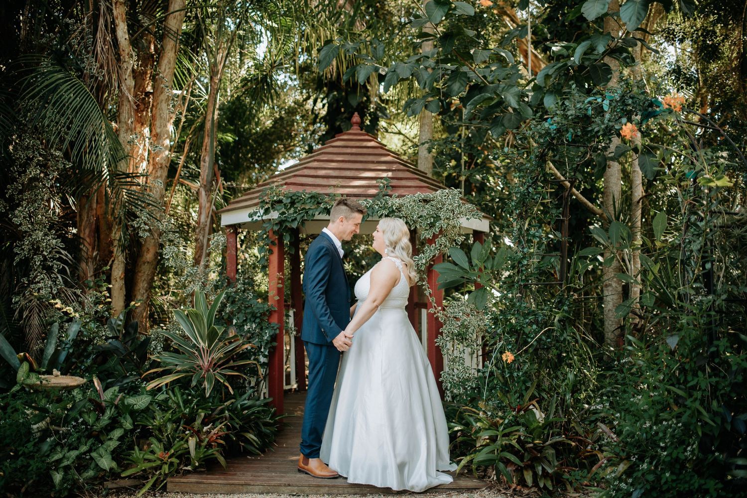 A couple stands together under a rustic wooden gazebo surrounded by lush tropical gardens.