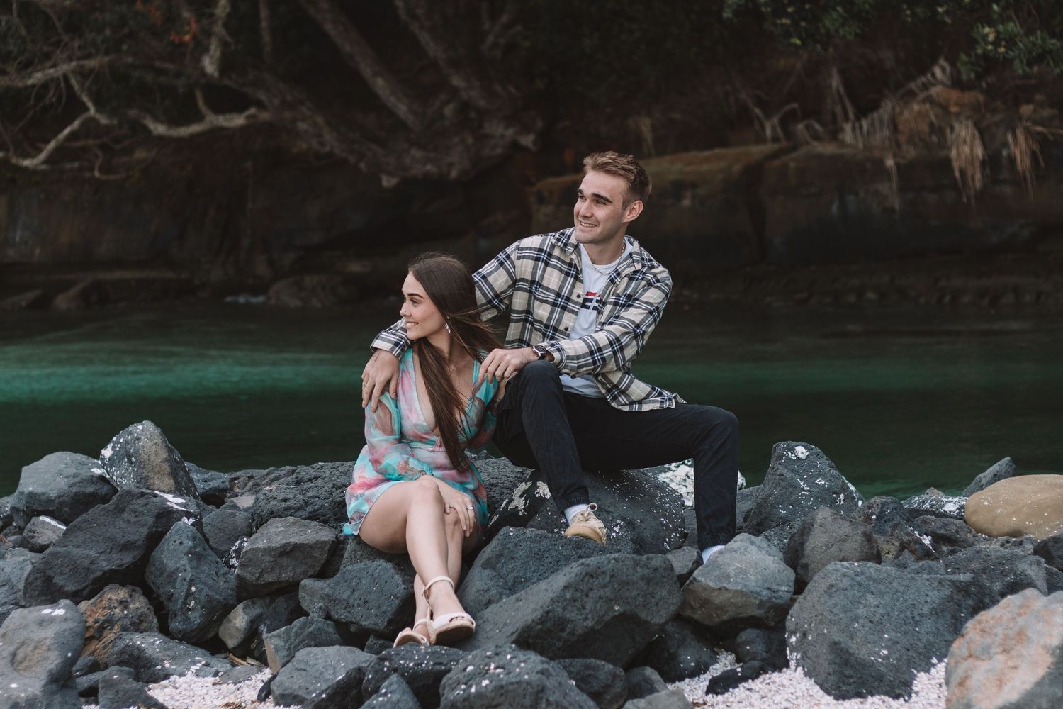 A couple sits casually on coastal rocks beside turquoise waters.
