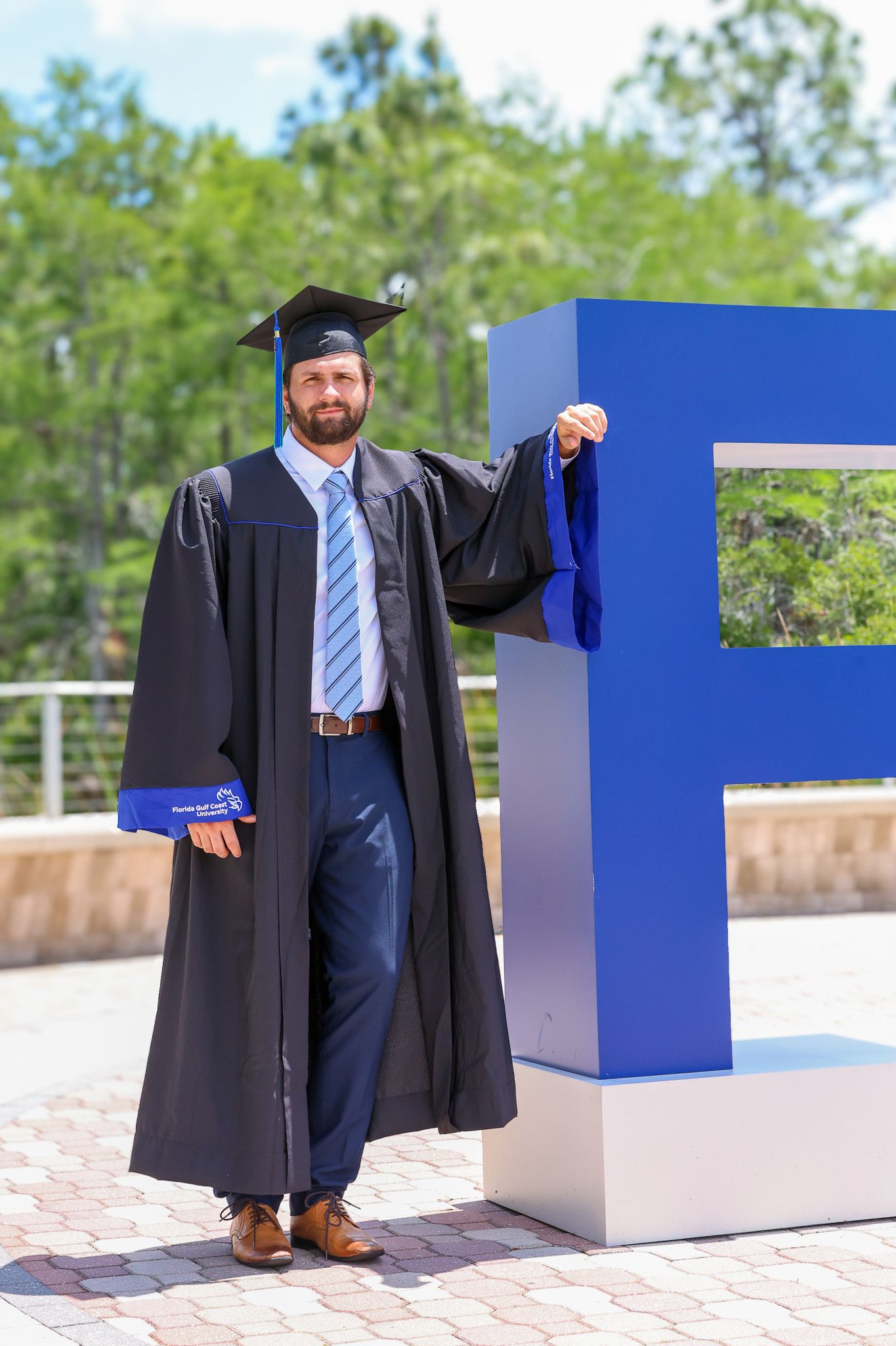 Graduate in cap and gown poses next to large blue F letter sculpture. Florida Gulf Coast University, Florida