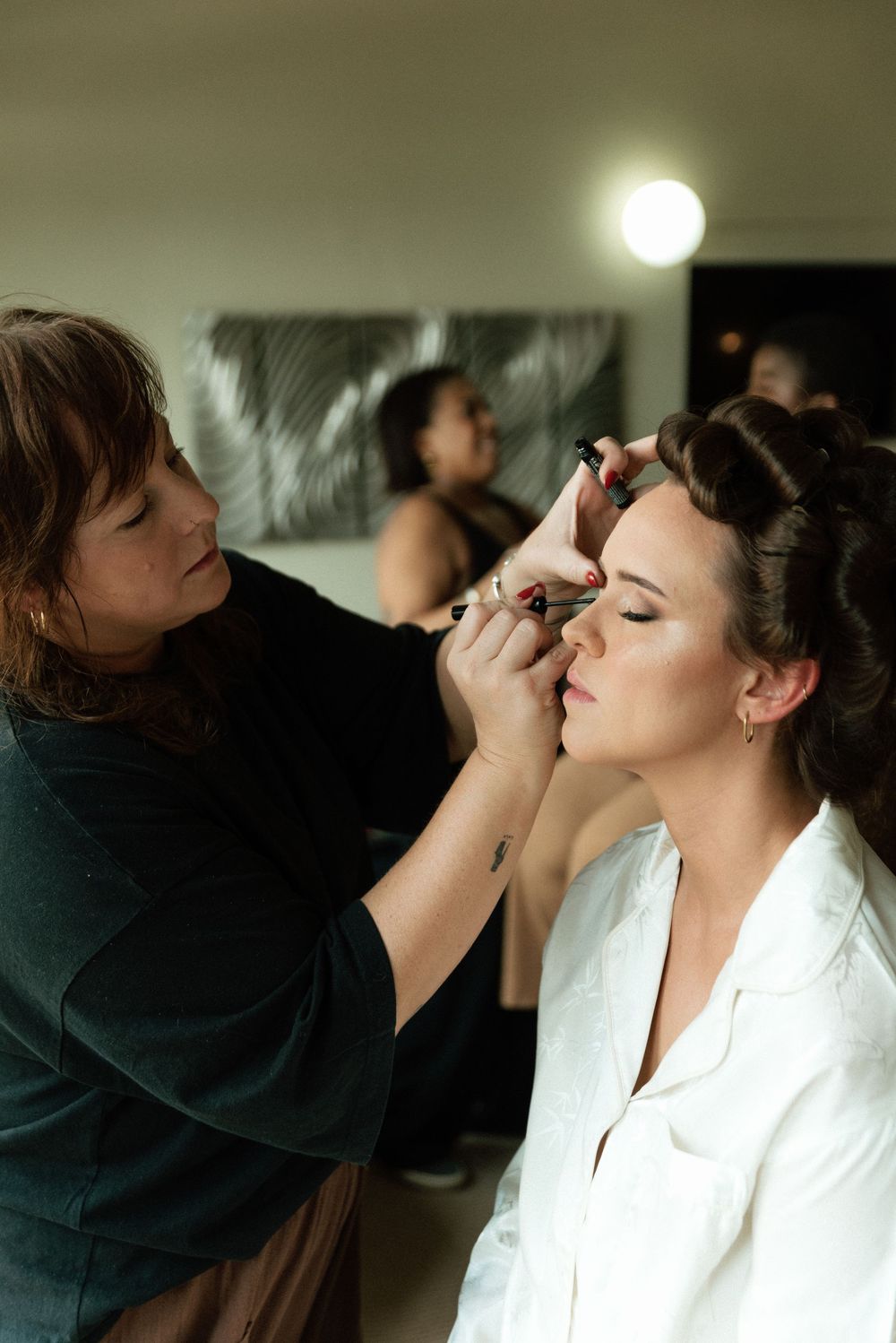 Makeup artist applying eye makeup to a bride with hair rollers, wearing a white robe, in a softly lit room.