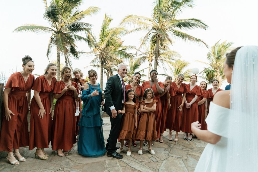 Bridesmaids in rust-colored dresses stand with groom near palm trees at tropical outdoor wedding ceremony.