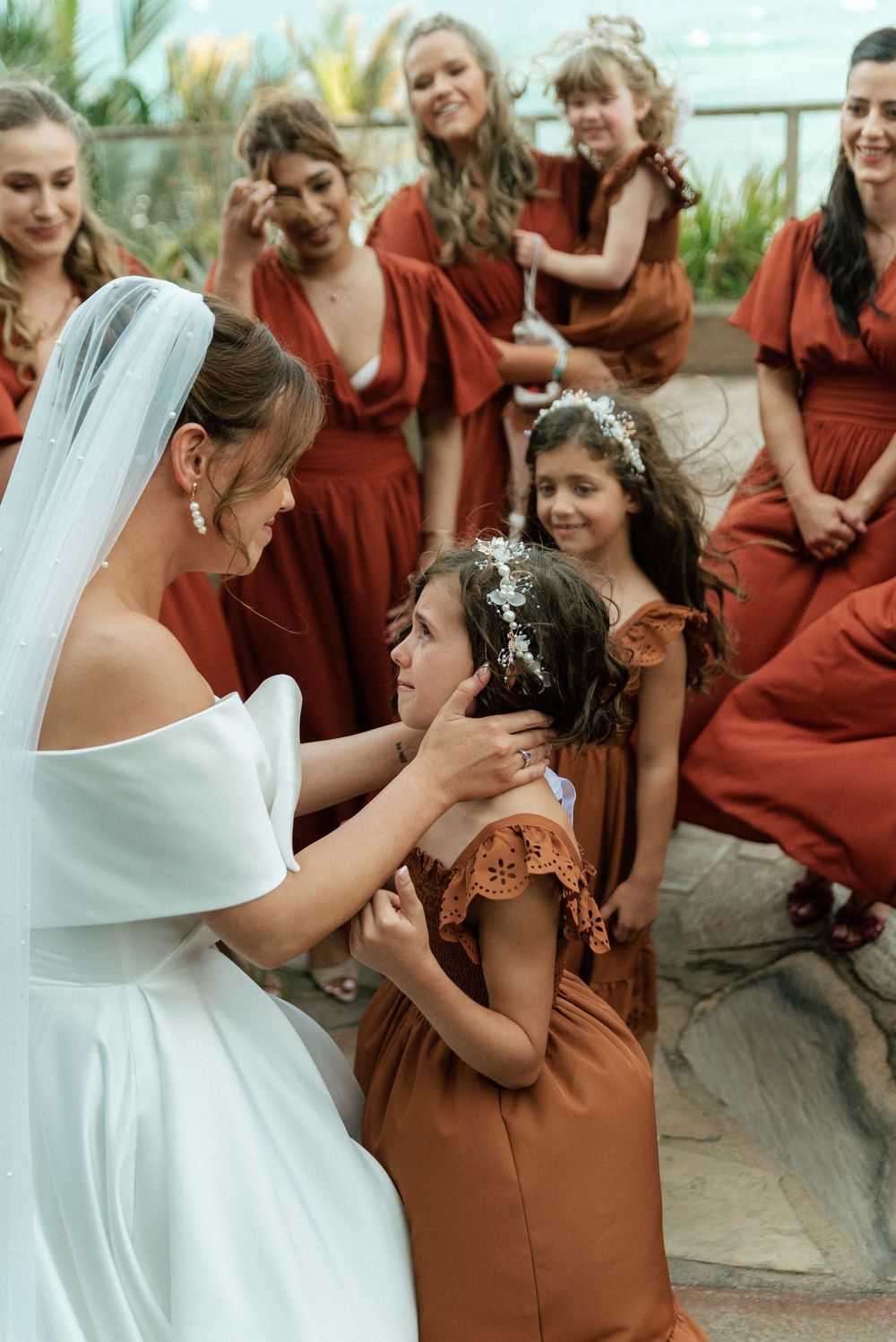 Bride in white gown adjusts flower girl's hair while bridesmaids in rust dresses look on at outdoor wedding.