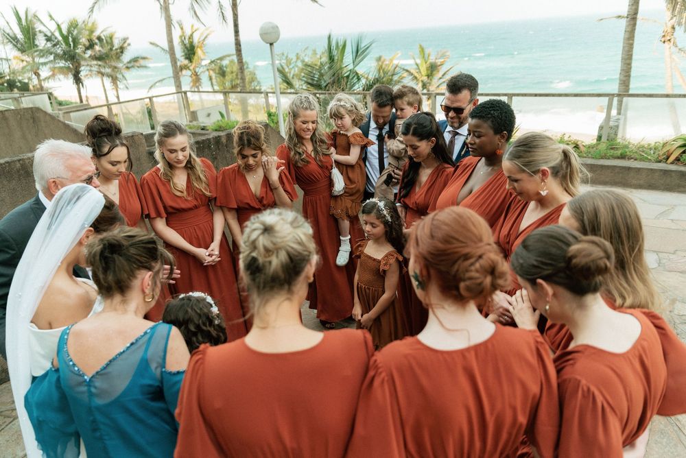 Wedding party in rust-colored bridesmaid dresses gathered in a circle outdoors near a beach venue.