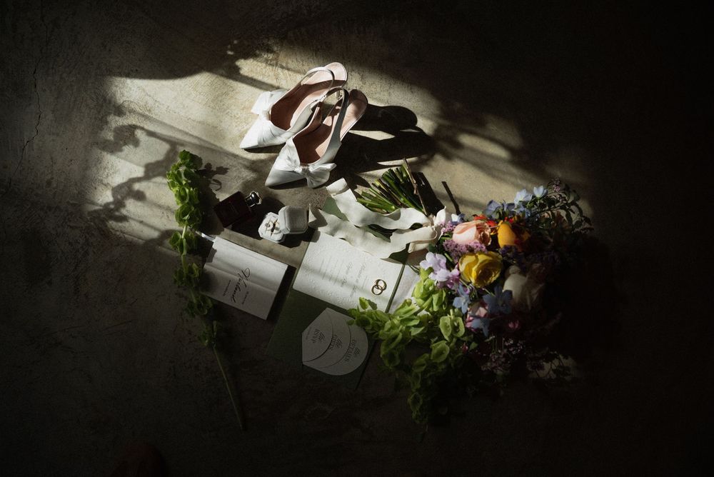 Flat lay of wedding shoes, floral bouquet, and beauty products arranged on a dark surface with dramatic lighting.