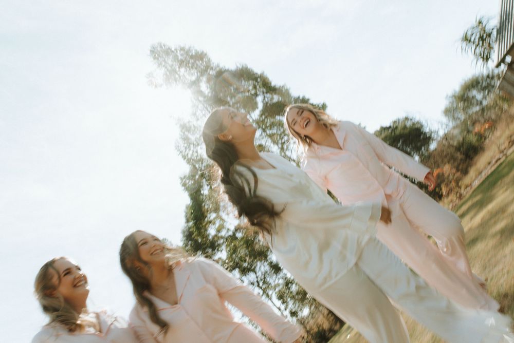 Four women in white outfits laughing and posing outdoors in bright sunlight with trees in the background.