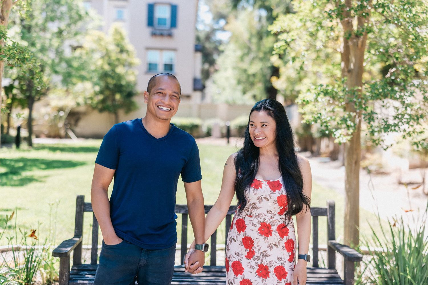 Two people pose together on a wooden deck in a sunny garden with trees and buildings in the background.