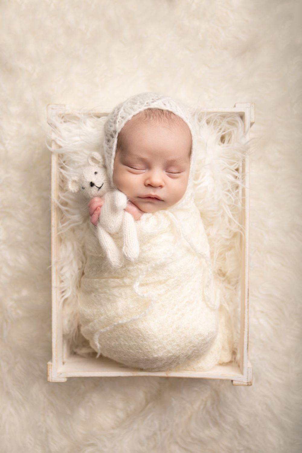 Sleeping newborn in white lace wrap with small stuffed toy in wooden crate.
