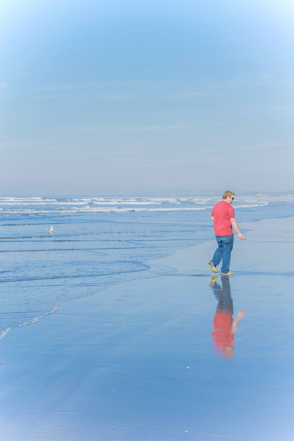 Person in coral shirt walking along misty ocean shoreline with reflection.