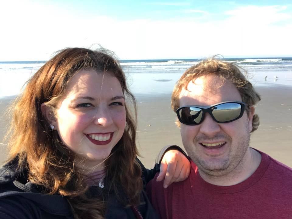 Couple taking selfie together on sunny beach with ocean waves behind them.