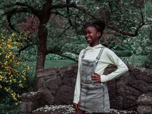 Person in overalls and cream sweater poses against stone wall with yellow flowers in background. Family Photography
