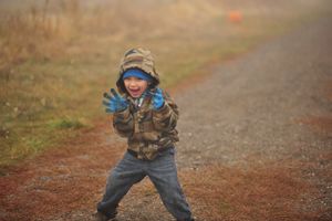 A child in winter clothing plays outdoors on a dirt path with expressive joy.