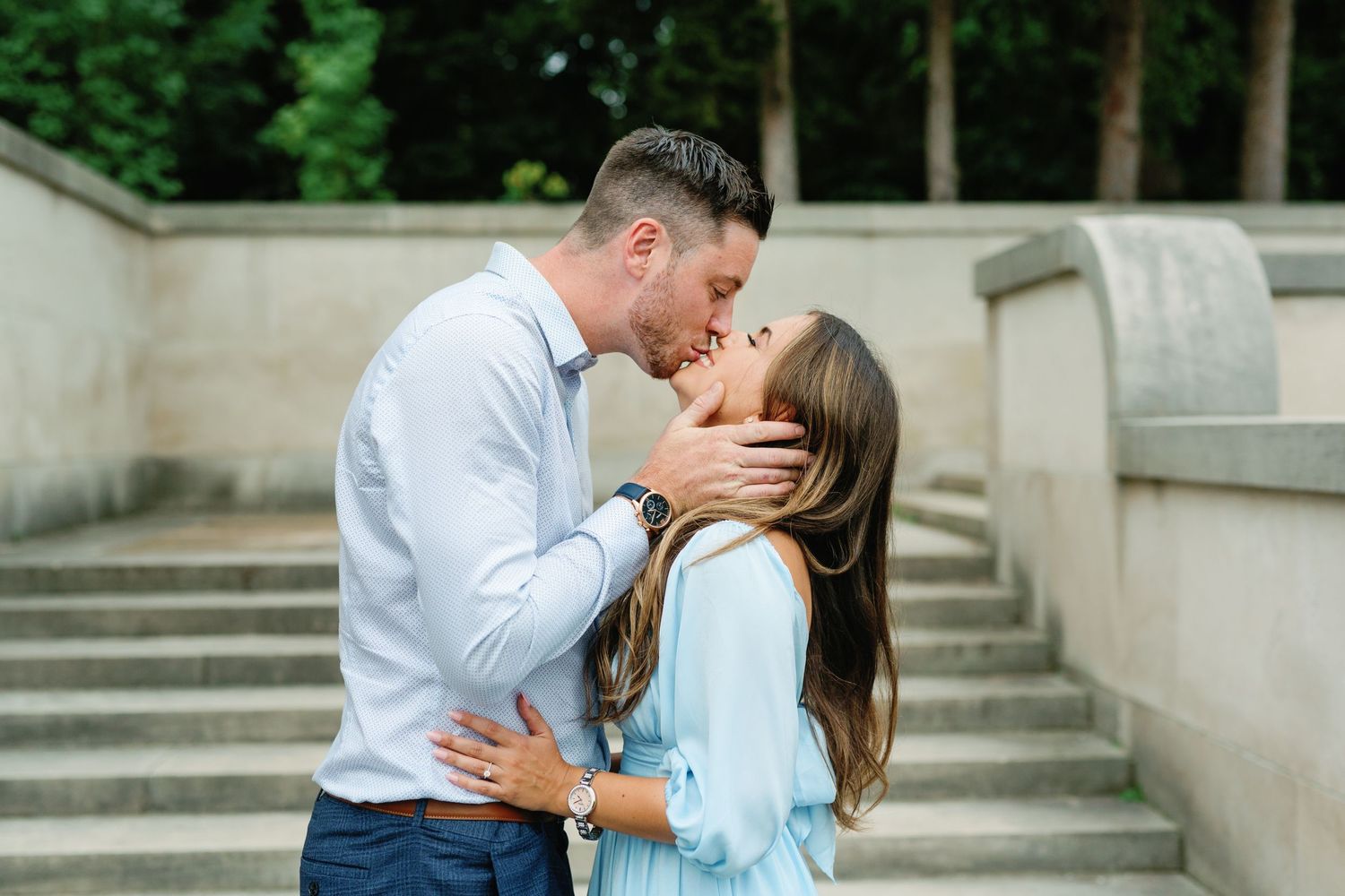 Couple sharing romantic kiss on stone steps in formal garden setting.