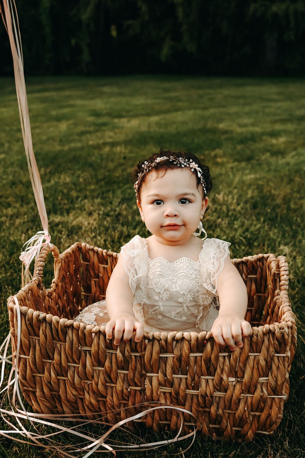 A baby wearing a white lace dress sitting in a woven basket outdoors on grass.