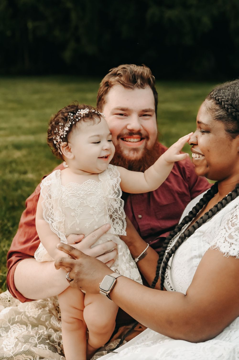 A joyful family moment outdoors with a baby in a white lace dress being held lovingly.
