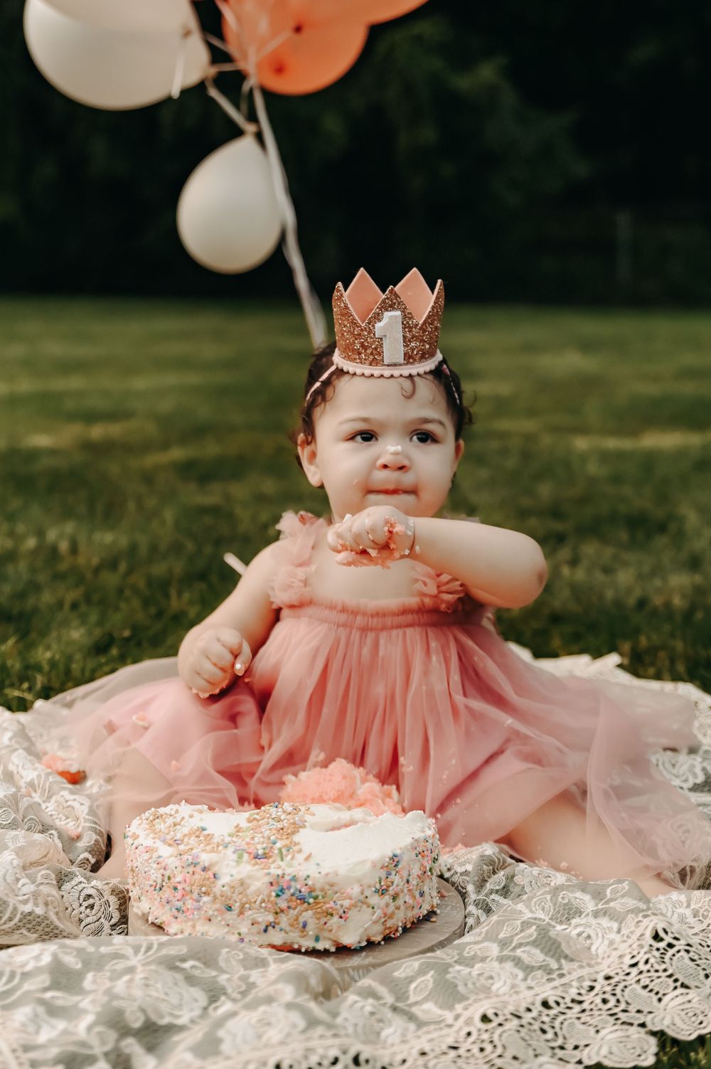 A baby in a pink tulle dress with a crown sitting on lace during an outdoor cake smash session.