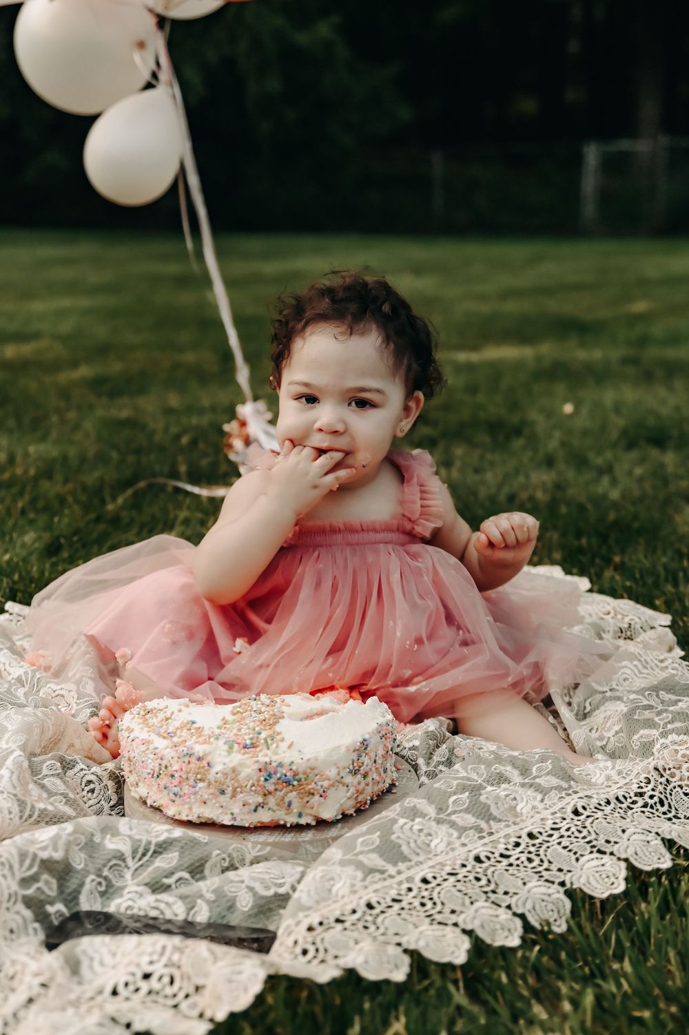 A baby in pink dress enjoying cake while sitting on white lace during an outdoor photoshoot.