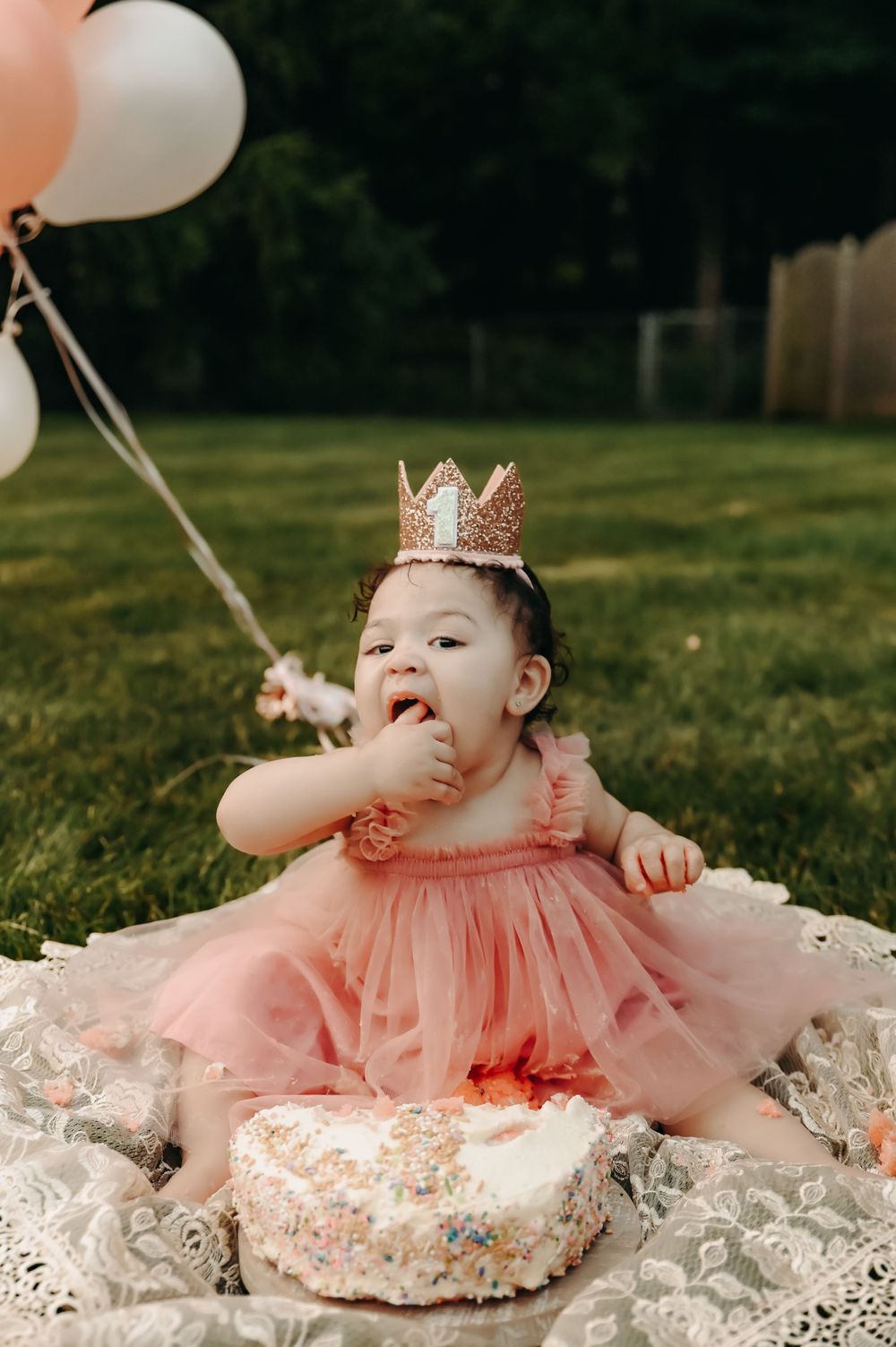 Baby wearing pink crown and dress smashes into birthday cake during cake smash photoshoot on grass.