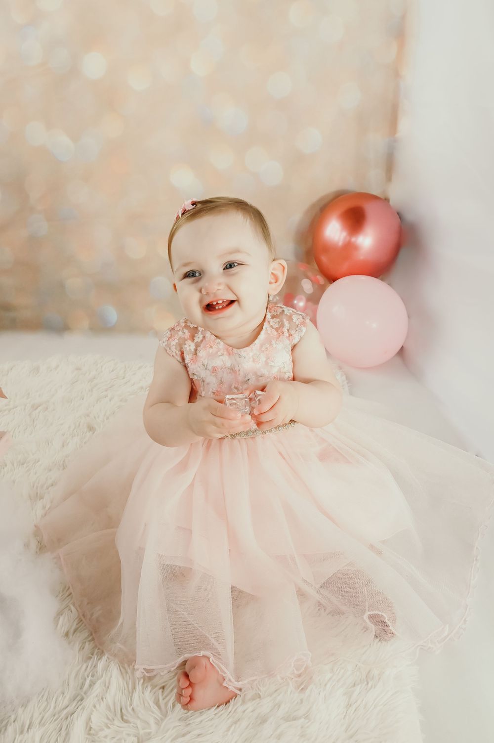 A baby in a pink tulle dress smiling and playing with pink balloons on a light background.