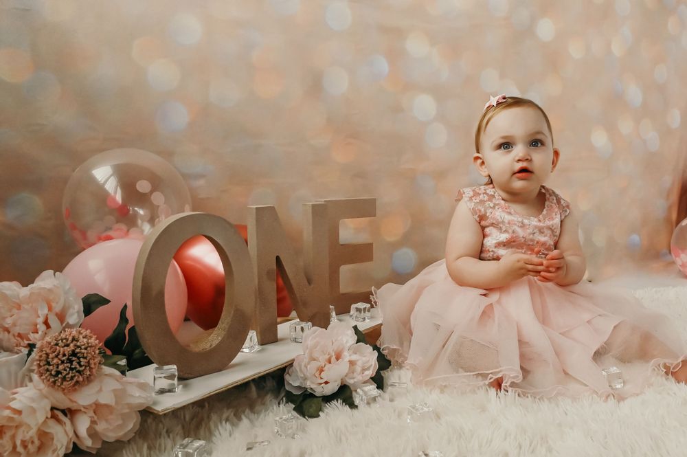 A baby in pink dress sitting among decorative elements and letters spelling 'ONE' for a first birthday shoot.