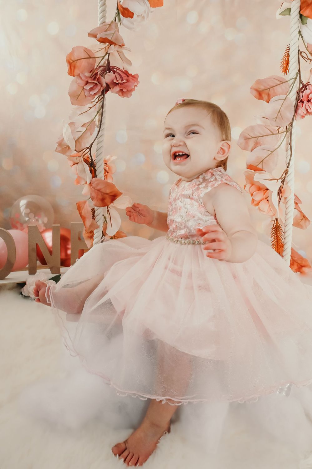 A baby wearing a pink dress laughing among coral flowers and white tulle decorations.
