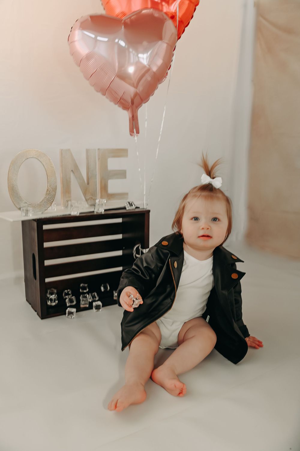 Baby in black outfit sits with pink heart balloons and ONE sign for first birthday photos.