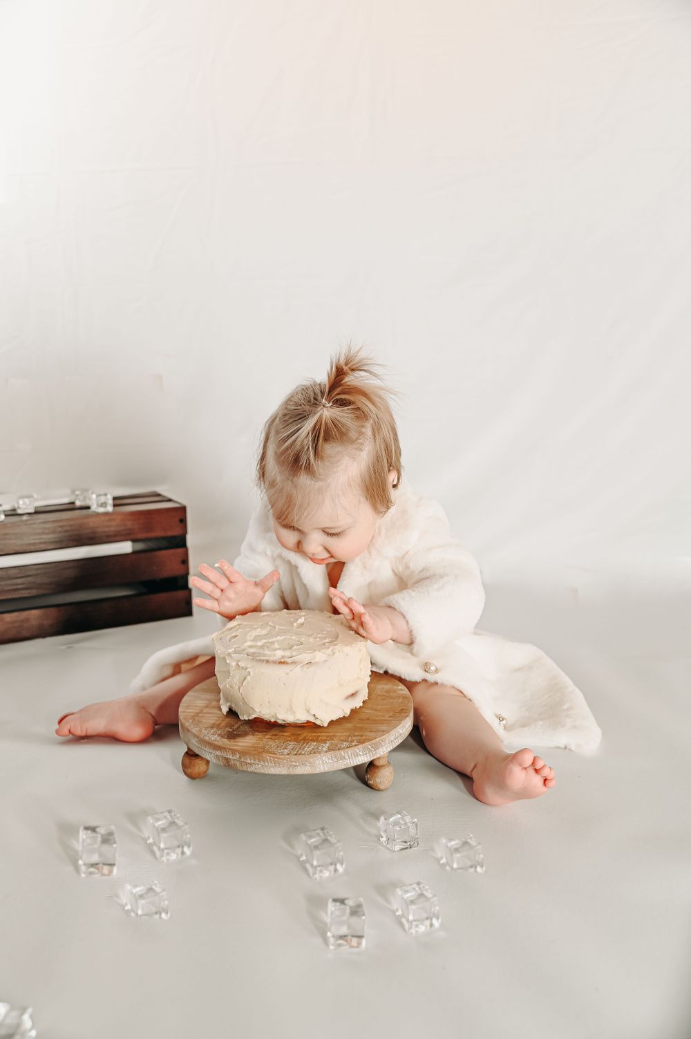 A baby in a pink dress lying on a white surface eating cake with decorative crystal elements nearby.