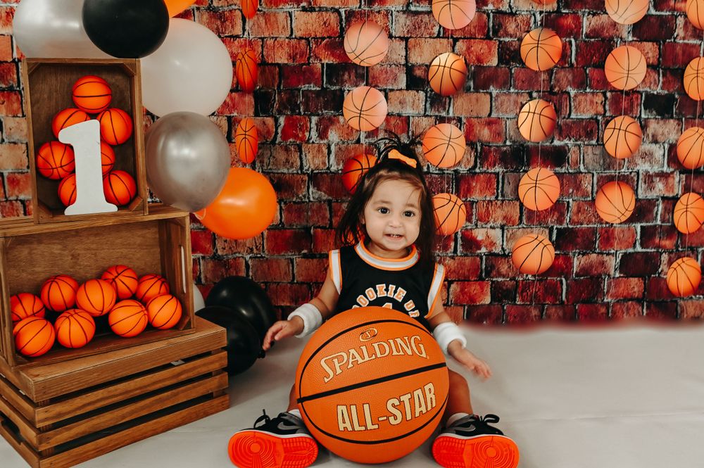 Basketball themed first birthday photoshoot with orange and white balloons against brick wall backdrop.