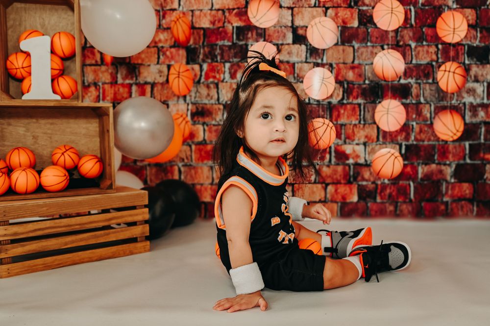 First birthday basketball themed photoshoot with child sitting on white floor against decorated brick wall.