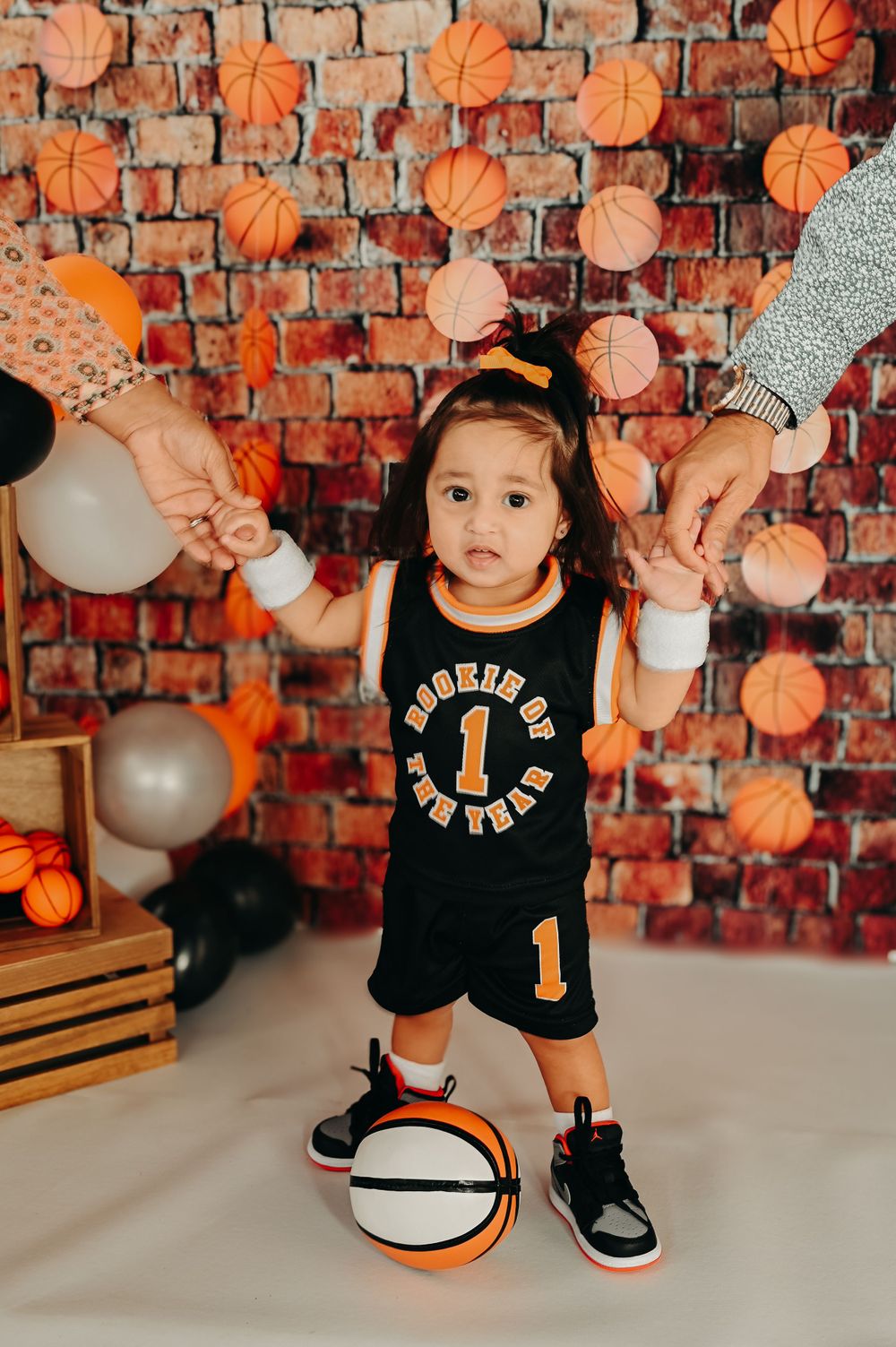 Child in basketball outfit standing with ball during first birthday photoshoot against decorated wall.