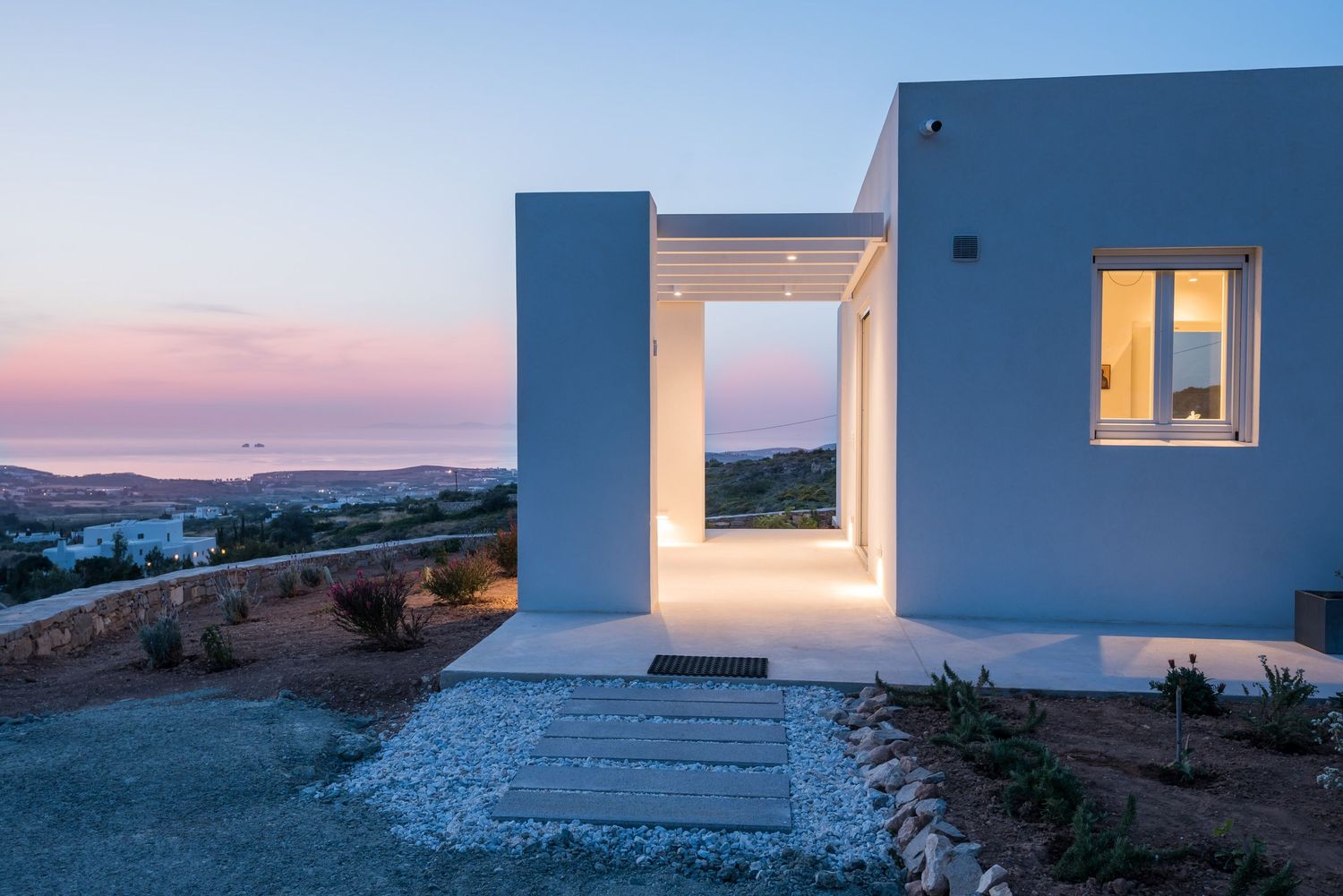 Modern white villa with illuminated entryway at sunset overlooking Mediterranean landscape in Greece.