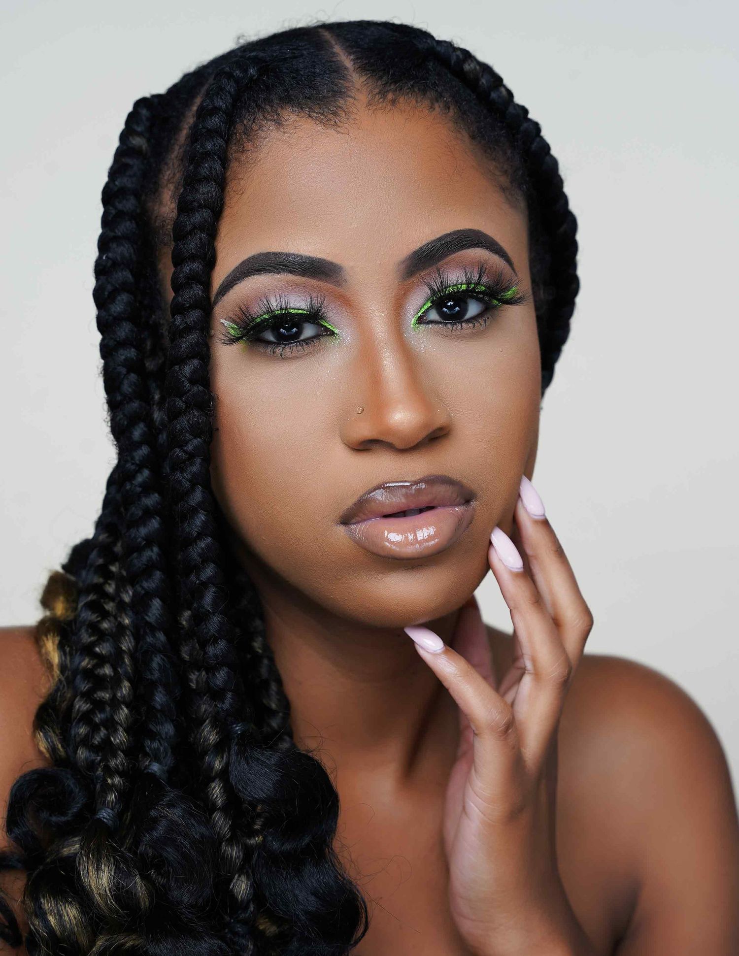 Portrait showing stunning eye makeup with green and pink eyeshadow, cornrow braids, and glossy nude lips against studio backdrop.