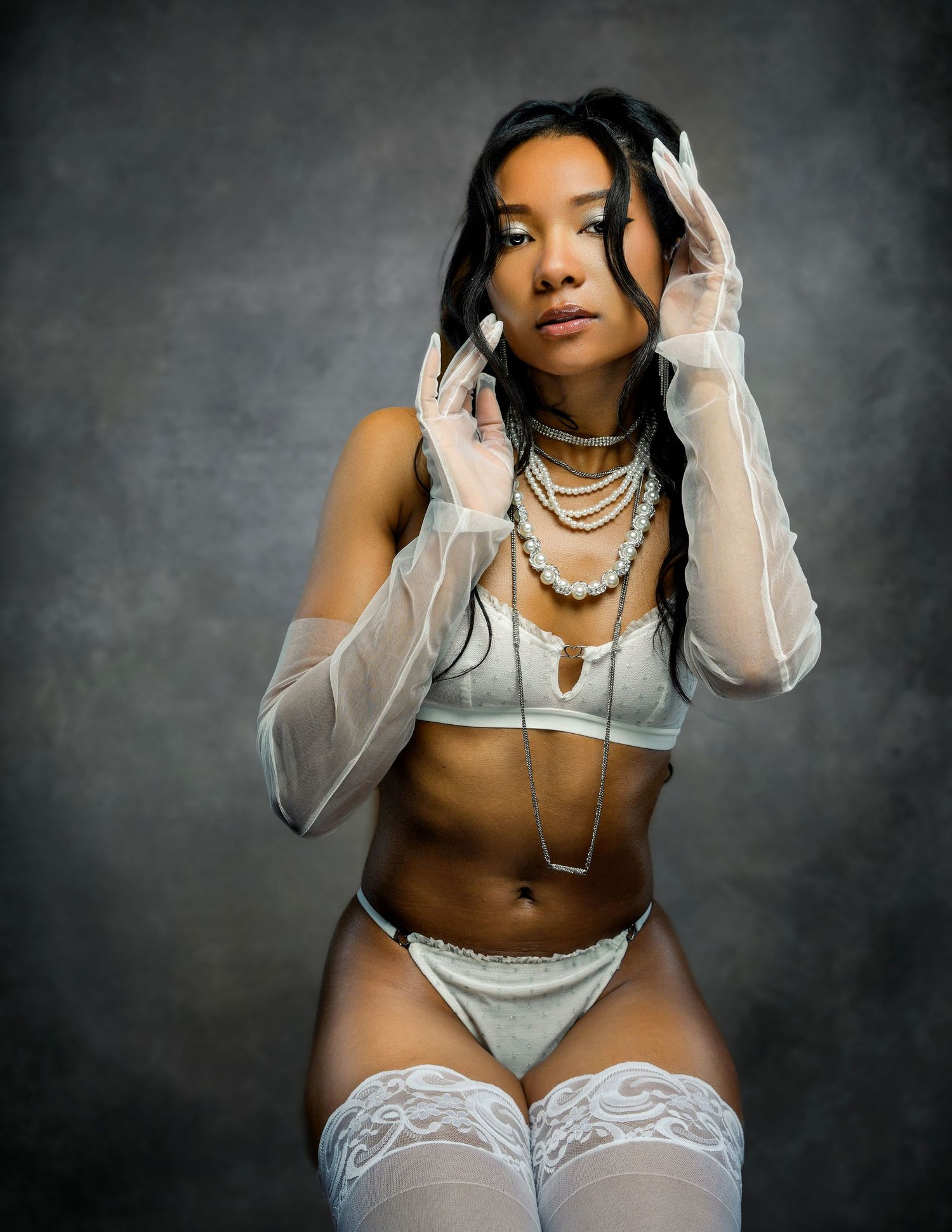 Woman in white lingerie and sheer gloves poses against dark backdrop with pearl necklaces.