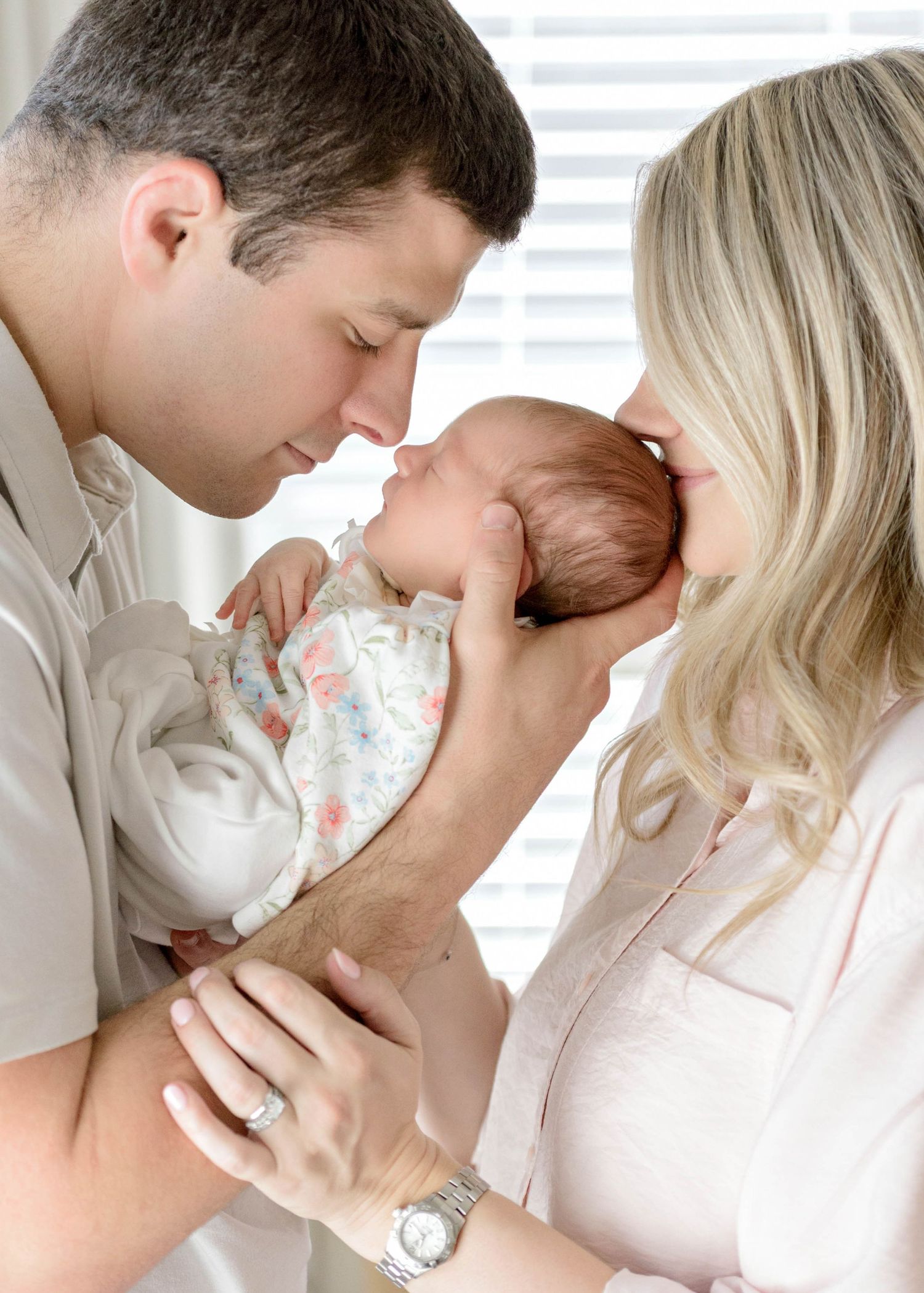Parents lovingly embrace their newborn in a tender family moment. Dad holding baby girl, nose to nose, and mom kissing top of her head.