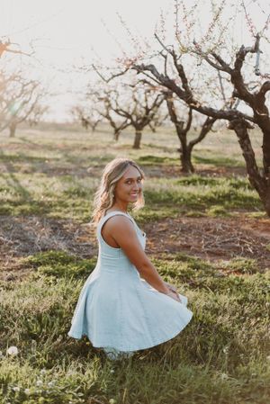 A light blue dress twirls in a rustic orchard setting during golden hour as sunlight filters through bare tree branches.