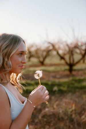 A dreamy sunset portrait shows a person holding a dandelion flower in an orchard during golden hour.