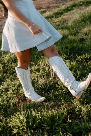 A person wearing a white dress and white cowboy boots standing on sunny grass in golden light.