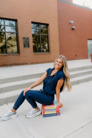 Student in casual denim outfit sitting on concrete steps with textbooks, wearing athletic sneakers next to a brick building.