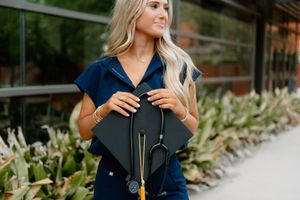 Person in navy blue top holding a black graduation cap and tassel against green foliage background.