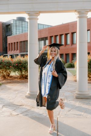 Graduate in black cap and gown poses with blue sash in front of brick university building with white columns.