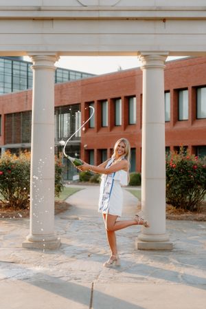 Someone in a white dress poses playfully between classical columns at sunset near a brick building with landscaping.