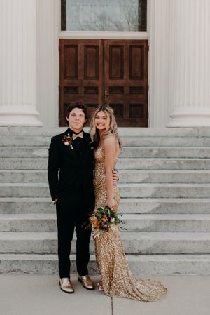 A couple in wedding attire poses on stone steps in front of a wooden door, wearing black suit and gold sequin gown.