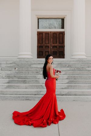 Elegant woman in flowing red mermaid gown holds bouquet while standing on grand white stone steps by wooden door.