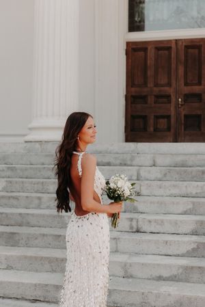 A bride in a white lace dress stands on marble steps holding a bouquet against classic white columns.