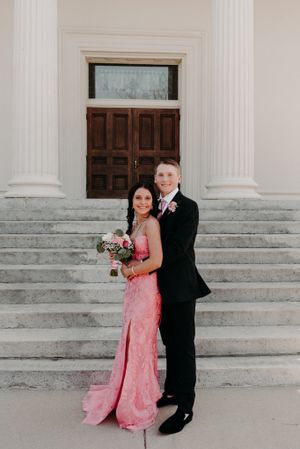 A couple poses together on grand white stone steps wearing formal attire, with a pink dress and black suit.