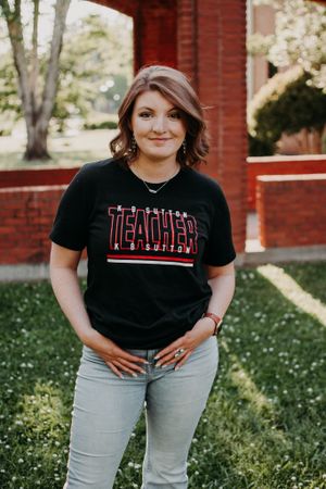 Woman wearing teacher t-shirt poses in front of brick building.