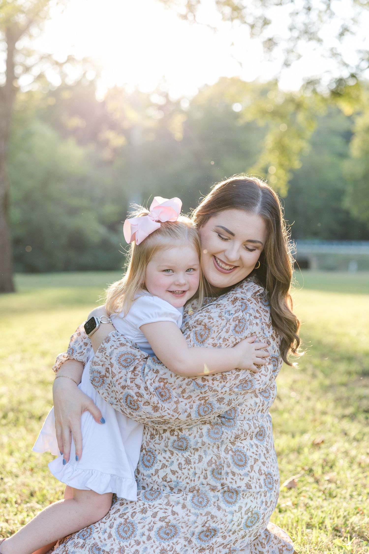 Mother and daughter share a joyful embrace while sitting in a sunlit grassy field at golden hour.