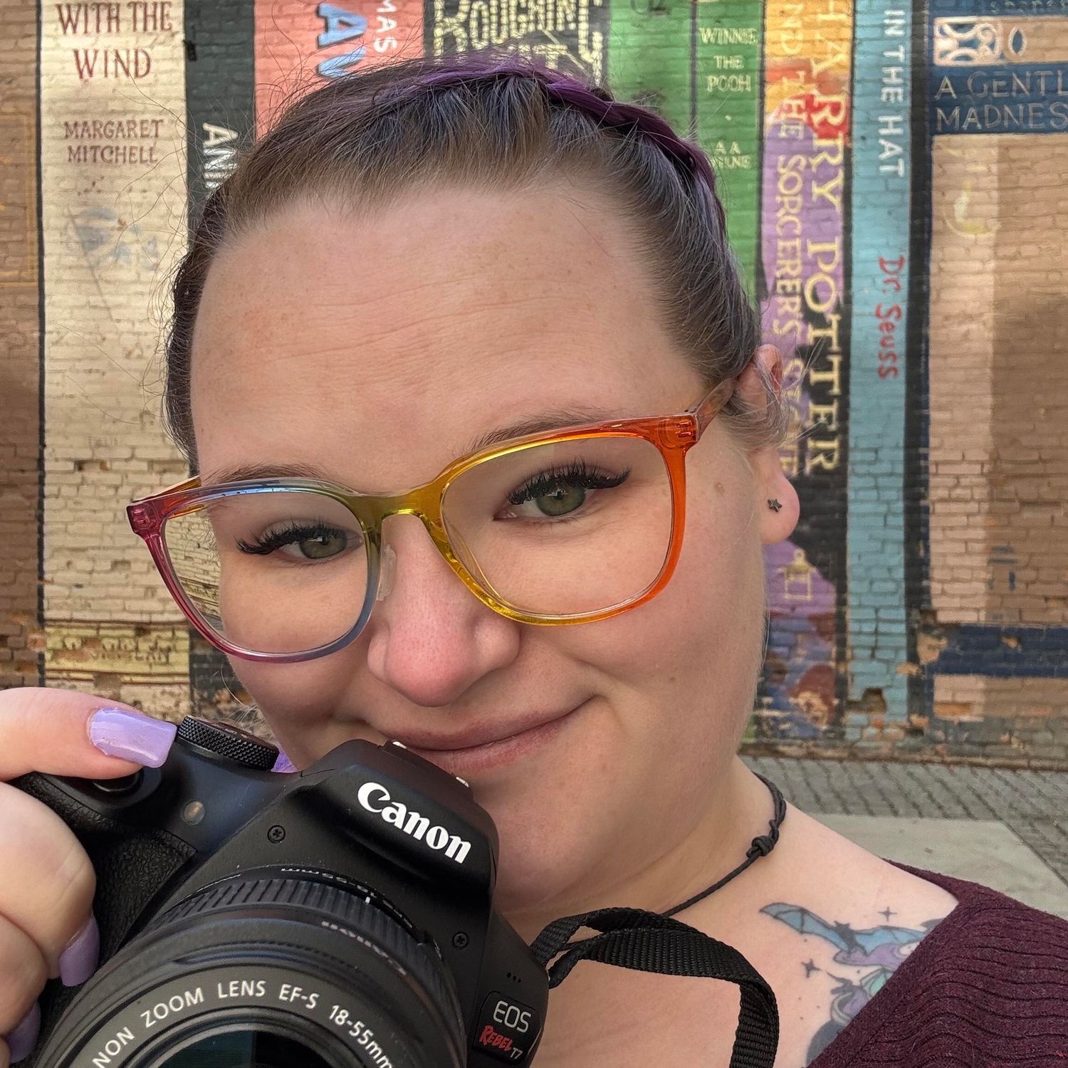 Photographer with vibrant purple hair holding a camera and wearing glasses against wooden background.