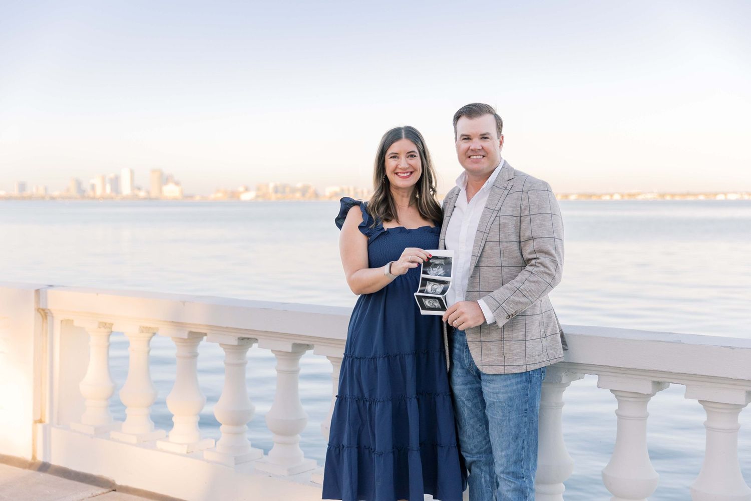 A couple poses at sunset on a waterfront balcony with white stone railings and city views in the background.
